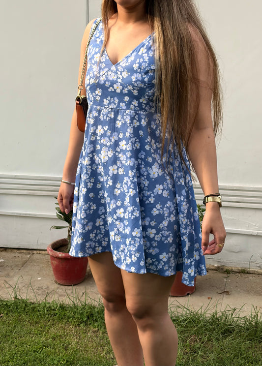 Person wearing a blue floral dress standing outdoors with a white wall and potted plants in the background.